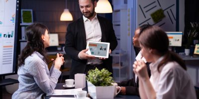 Entrepreneur businessman showing company strategy using tablet for corporate presentation working overtime in meeting office room late at night. Diverse multi-ethnic teamwork brainstorming ideas.