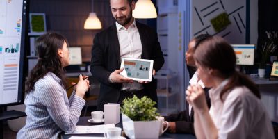 Entrepreneur businessman showing company strategy using tablet for corporate presentation working overtime in meeting office room late at night. Diverse multi-ethnic teamwork brainstorming ideas.
