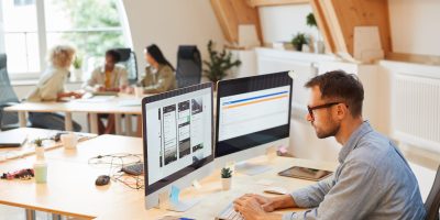 Serious programmer sitting at his workplace using computers in his work he developing new software at office with his colleagues in the background